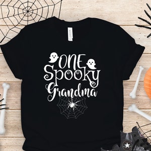 May include: Black t-shirt with white text that reads "One Spooky Grandma" with two ghost images and a spider web with a spider in the center.