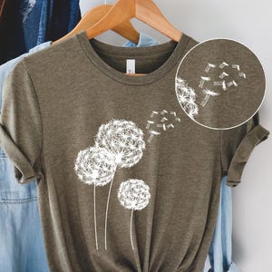 May include: Olive green t-shirt featuring a white dandelion design. The graphic shows three dandelion seed heads with seeds blowing away. The shirt is hanging on a wooden hanger with other clothing items in the background.