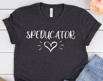 May include: A dark gray t-shirt with the text "SPEDUCATOR" and a white heart design. The shirt is laid out on a white wooden surface with a pair of brown sandals and blue jeans.