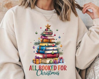 May include: Beige sweatshirt with a Christmas tree design made of books. The tree is decorated with ornaments and topped with a star. The text "ALL BOOKED FOR Christmas" is printed below the tree in red and green.