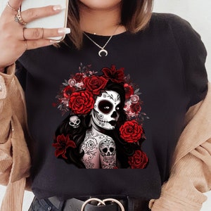 May include: Black t-shirt with a colorful illustration of a woman with a sugar skull face and red roses. The woman has tattoos and is wearing a flower crown.