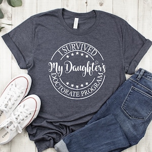 May include: A dark heather gray t-shirt with a white circle graphic that says "I Survived My Daughter's Doctorate Program".