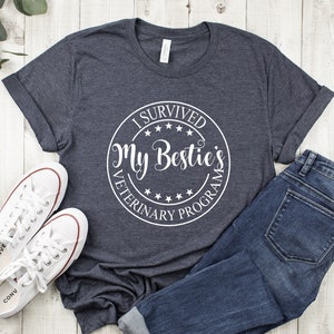 May include: A dark heather grey t-shirt with a white circle graphic that says "I Survived My Bestie's Veterinary Program".