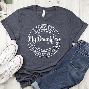 May include: A dark heather grey t-shirt with a white circle graphic that says "I Survived My Daughter's Veterinary Program".