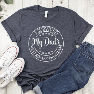 May include: A dark gray t-shirt with a white circle graphic that says "I Survived My Dad's Veterinary Program". The shirt is laid out on a white wooden surface with a pair of white sneakers and blue jeans.
