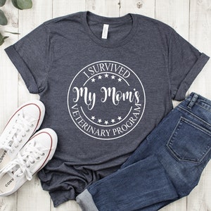May include: A dark heather grey t-shirt with a white circle graphic that says "I Survived My Mom's Veterinary Program". The graphic has five stars around the outside of the circle.