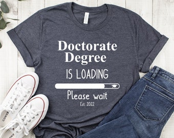 May include: A dark gray t-shirt with white text that reads "Doctorate Degree is Loading. Please wait. Est. 2022". The text is in the style of a loading bar with a graduation cap at the end.