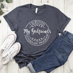 May include: A dark gray t-shirt with a white circle graphic that says "I Survived My Girlfriend's Veterinary Program".