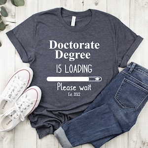 May include: A dark gray t-shirt with white text that reads "Doctorate Degree is Loading. Please wait. Est. 2022". The text is in the style of a loading bar with a graduation cap at the end.