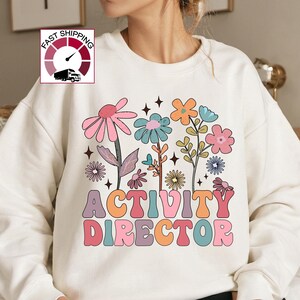 May include: Cream-colored sweatshirt featuring a colorful floral design and the text "ACTIVITY DIRECTOR" in a retro font. The flowers include pink, blue, orange, and yellow blooms. The sweatshirt is a casual style.