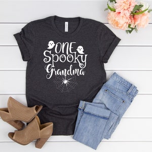 May include: A dark gray t-shirt with the text "One Spooky Grandma" in white with a ghost and spider graphic. The shirt is laying on a white wooden surface with a pair of brown shoes and blue jeans.