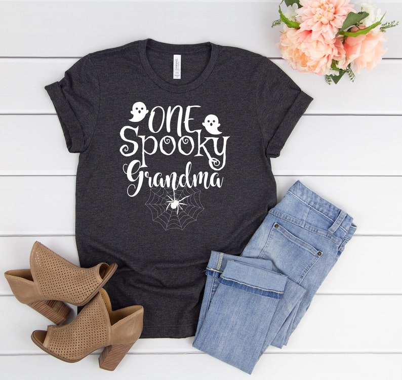 May include: A dark gray t-shirt with the text "One Spooky Grandma" in white with a ghost and spider graphic. The shirt is laying on a white wooden surface with a pair of brown shoes and blue jeans.