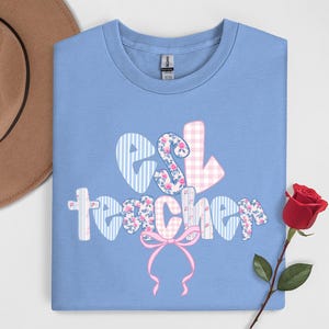 May include: A light blue t-shirt with the words "ESL teacher" in a decorative font. The letters are patterned with stripes, floral prints, and gingham. A pink bow is below the word "teacher". A brown hat is in the background.