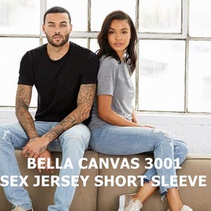 May include: Two people are wearing Bella Canvas 3001 Unisex Jersey Short Sleeve Tees. The person on the left is wearing a black tee and the person on the right is wearing a gray tee. Both are wearing blue jeans.