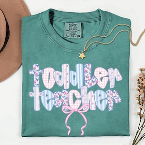 May include: Teal Comfort Colors t-shirt with the words "Toddler Teacher" in a floral, gingham, and striped design. A gold star pendant necklace and a brown hat are also visible.