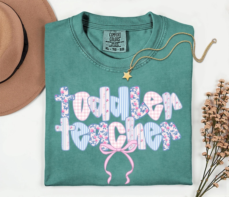 May include: Teal Comfort Colors t-shirt with the words "Toddler Teacher" in a floral, gingham, and striped design. A gold star pendant necklace and a brown hat are also visible.