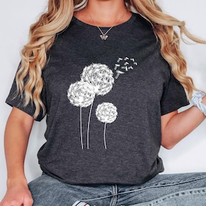 May include: Dark gray t-shirt featuring a white dandelion design. The graphic shows three dandelion seed heads with some seeds blowing away. The shirt has short sleeves and a crew neck.
