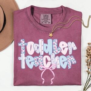 May include: A maroon Comfort Colors t-shirt with the words "Toddler Teacher" in a floral, gingham, and striped design. A gold necklace with a star pendant and a brown hat are also visible.