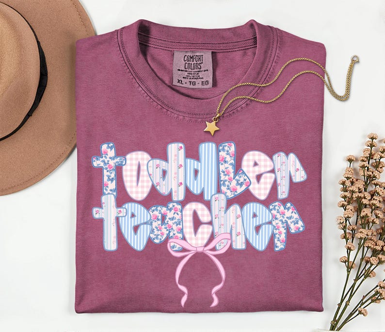 May include: A maroon Comfort Colors t-shirt with the words "Toddler Teacher" in a floral, gingham, and striped design. A gold necklace with a star pendant and a brown hat are also visible.