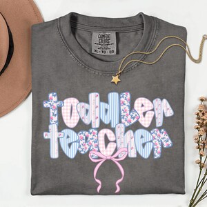 May include: A gray Comfort Colors t-shirt with the words "Toddler Teacher" in a bubble font. The letters are decorated with pink floral, blue striped, and pink gingham patterns. A gold star pendant necklace and a pink bow are also visible.