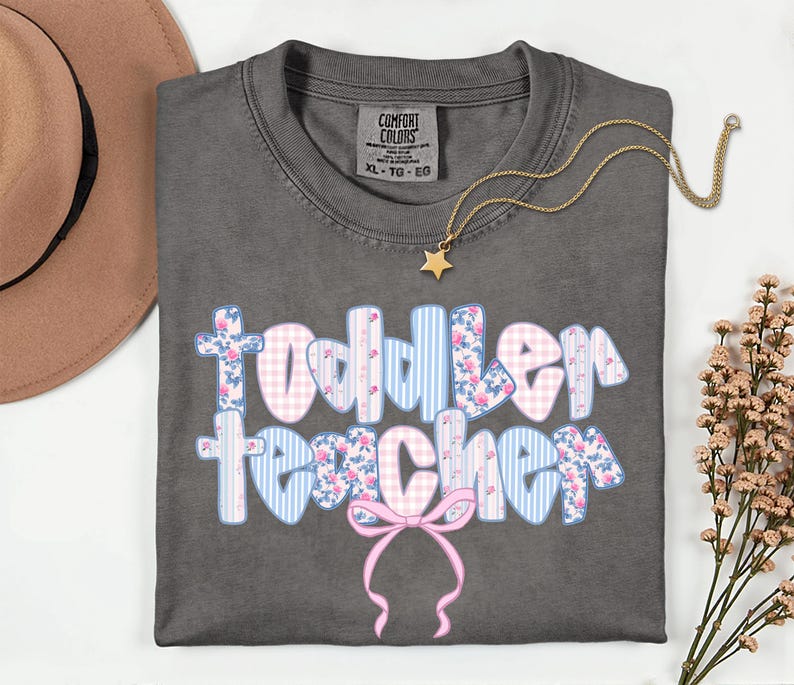May include: A gray Comfort Colors t-shirt with the words "Toddler Teacher" in a bubble font. The letters are decorated with pink floral, blue striped, and pink gingham patterns. A gold star pendant necklace and a pink bow are also visible.