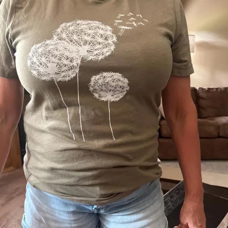 May include: Olive green t-shirt featuring a white dandelion design. The graphic includes three large dandelion seed heads and several seeds blowing away. The shirt is made of a soft material and is suitable for casual wear.