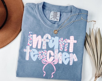 Könnte beinhalten: Ein blaues Comfort Colors T-Shirt mit den Worten "Infant Teacher" in einem Patchwork-Design. Die Buchstaben weisen florale, Vichy- und Streifenmuster in Rosa und Blau auf. Eine goldene Halskette und ein brauner Hut sind ebenfalls sichtbar.