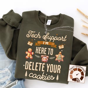 May include: An olive green sweatshirt with the text "Tech Support I'm Here To Delete Your Cookies". The design includes a gingerbread man, a bell, a Christmas tree, and gift boxes. A silver chain is draped across the top.