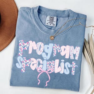 May include: A blue short-sleeved t-shirt with a crew neckline featuring the text "PROGRAM SPECIALIST" in a multi-patterned font with floral, gingham, and striped designs in pink and blue, accented by a pink bow graphic below the text.