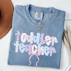 May include: A blue Comfort Colors t-shirt with the words "Toddler Teacher" in a floral and gingham pattern. A gold necklace with a pendant is on the shirt. A pink bow is at the bottom of the text.