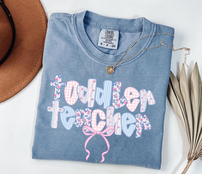 May include: A blue Comfort Colors t-shirt with the words "Toddler Teacher" in a floral and gingham pattern. A gold necklace with a pendant is on the shirt. A pink bow is at the bottom of the text.