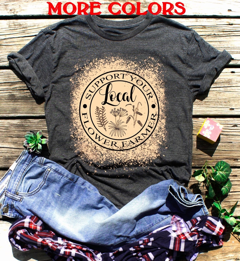 Puede incluir: Una camiseta gris oscuro con un dise&ntilde;o salpicado de lej&iacute;a que presenta un c&iacute;rculo con el texto "Apoya a tu agricultor local de flores" y una ilustraci&oacute;n floral.