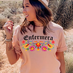 May include: A pink t-shirt with the word "Enfermera" in black lettering and a colorful floral design. The shirt has short sleeves and a crew neck.