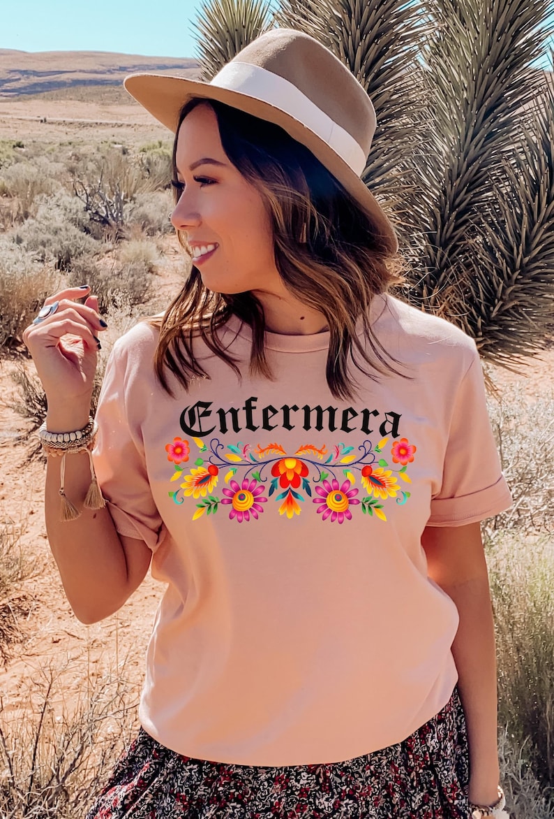 May include: A pink t-shirt with the word "Enfermera" in black lettering and a colorful floral design. The shirt has short sleeves and a crew neck.