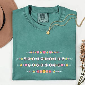 May include: Teal Comfort Colors t-shirt with the text "ACTIVITIES DIRECTOR" in a colorful, whimsical font. The shirt features decorative floral and heart accents. A gold star pendant necklace rests on the shirt.