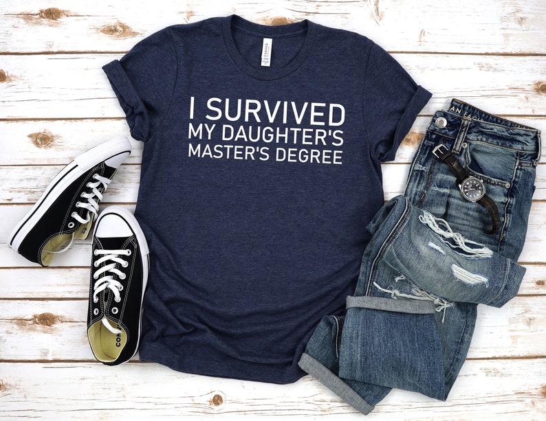 Peut inclure: Un t-shirt bleu marine avec un texte blanc qui dit "J'ai surv&eacute;cu au master de ma fille". Le t-shirt est pr&eacute;sent&eacute; sur un fond en bois blanc avec une paire de baskets noires et un jean bleu.