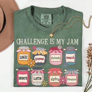 May include: Green t-shirt with the text "CHALLENGE IS MY JAM" and colorful jars labeled with subjects like Logic, Grammar, and Debate. A gold necklace with a star pendant hangs on the shirt.