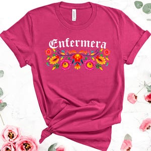 May include: Pink t-shirt with the word "Enfermera" in white lettering with a floral design underneath. The flowers are in shades of red, yellow, orange, and pink.