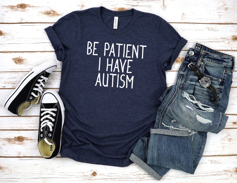 Op de afbeelding: Een marineblauw t-shirt met witte tekst die "Be Patient I Have Autism" luidt. Het shirt ligt plat op een wit houten oppervlak met een paar zwarte sneakers en een blauwe spijkerbroek.