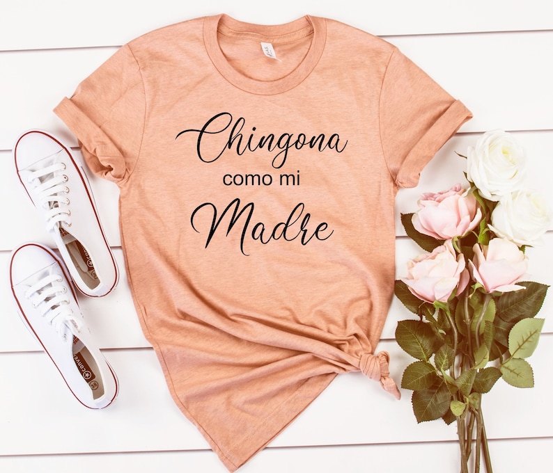 May include: A peach-colored t-shirt with the text "Chingona como mi Madre" in black cursive lettering. The shirt is knotted at the bottom and is photographed on a white wooden surface with white sneakers and pink roses.