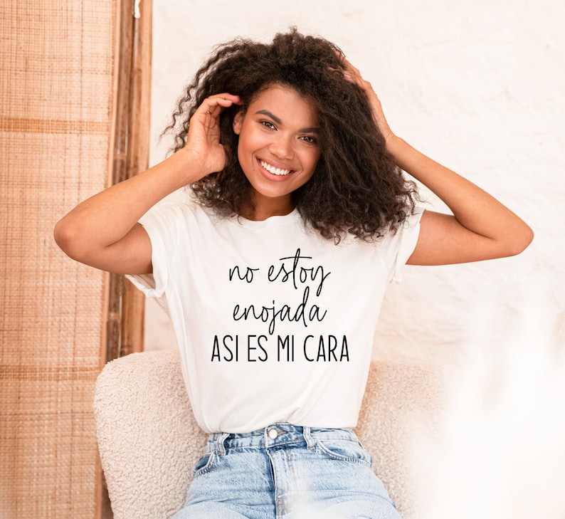 Op de afbeelding: Een wit T-shirt met zwarte tekst die "no estoy enojada asi es mi cara" zegt. De persoon die het shirt draagt ​​lacht en heeft krullend haar.