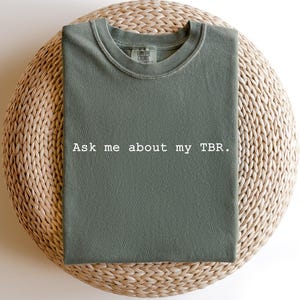 May include: Sage green t-shirt with the text "Ask me about my TBR" in white. The shirt is made of a soft material and has a crew neck. The shirt is displayed on a woven, round surface.