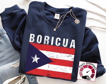 May include: Navy blue sweatshirt with the word "BORICUA" in white block letters above a distressed Puerto Rican flag graphic. A silver necklace with a pendant rests on the sweatshirt. The sweatshirt is folded on top of blue jeans and white sneakers.