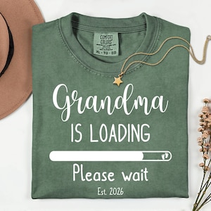 May include: Green t-shirt with the text "Grandma is Loading" in white script. The design includes a loading bar graphic and the words "Please wait" and "Est. 2026". A gold necklace with a star pendant is draped on the shirt.