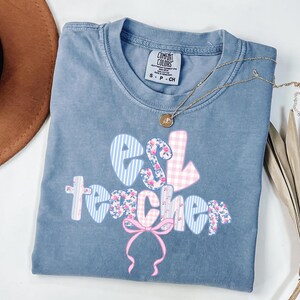 May include: A blue Comfort Colors t-shirt with the words "ESL teacher" in a mix of patterns and colors, including blue stripes, pink gingham, and floral prints. A gold necklace with a pendant is draped across the shirt.