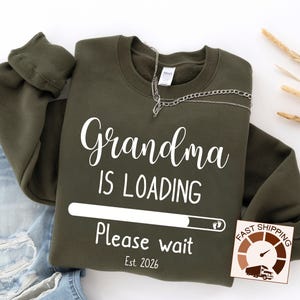 May include: An olive green sweatshirt with the text "Grandma IS LOADING Please wait Est. 2026" in white script. A loading bar graphic and a baby footprint are also featured. A silver chain necklace is draped across the top.