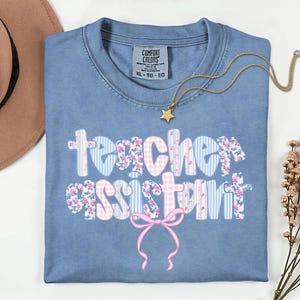 May include: A blue Comfort Colors t-shirt with the words "Teacher Assistant" in a floral and striped design. A gold necklace with a star pendant is draped across the shirt. A brown hat is visible in the upper left corner.