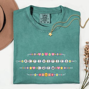 May include: Teal Comfort Colors t-shirt with the text "ACTIVITIES CREW" in a decorative font, adorned with colorful flower, heart, and butterfly graphics. A gold star pendant necklace rests on the shirt. A brown hat is in the background.