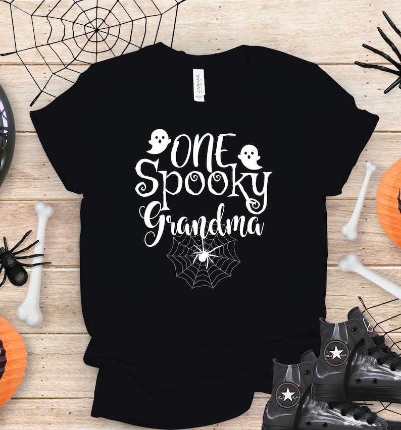 May include: Black t-shirt with white text that reads "One Spooky Grandma" with two ghost images and a spider web with a spider in the center.