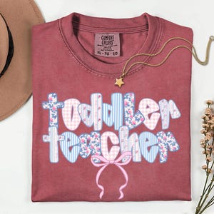 May include: A maroon Comfort Colors t-shirt with the words "Toddler Teacher" in a floral, gingham, and striped design. A gold star pendant necklace and a tan hat are also visible.